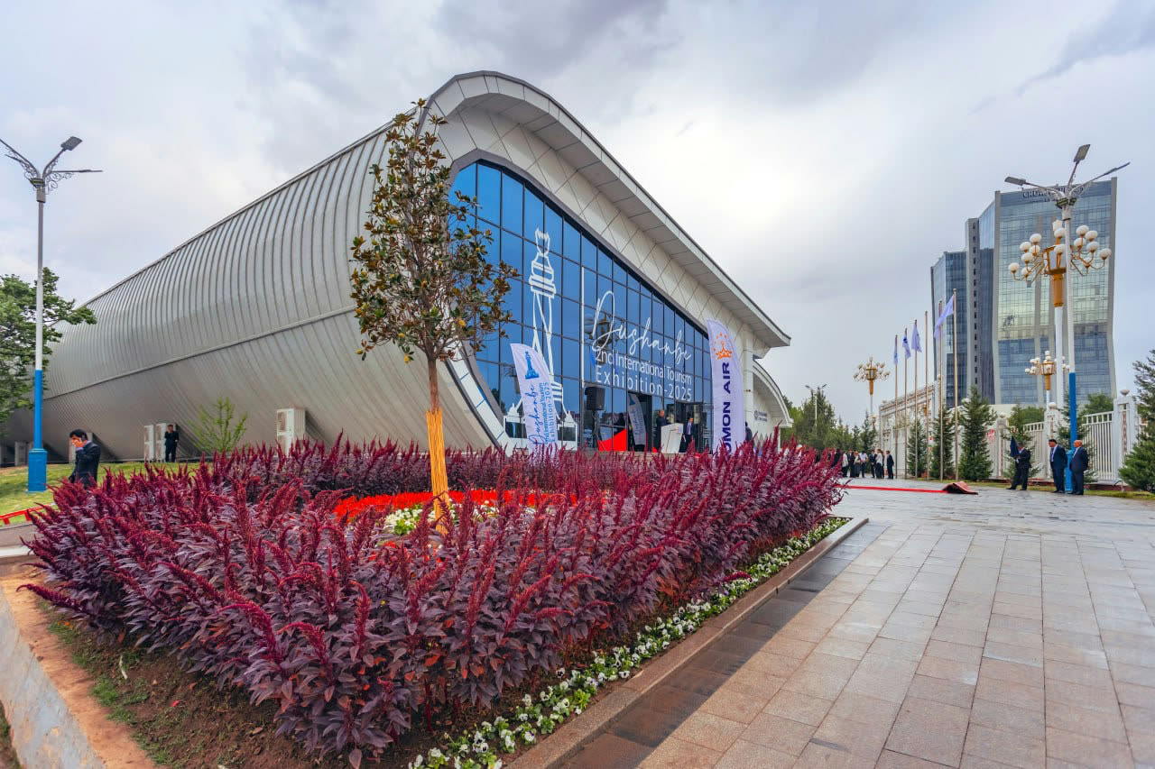 The Expo Center’s curved glass facade, its window lettered “2nd International Tourism Exhibition-2025”, behind a bed of dark red flowers