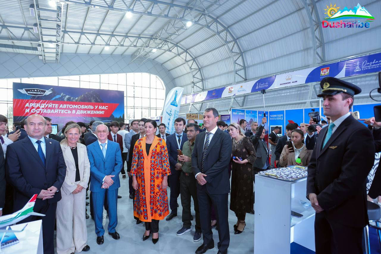 Visitors and officials inside the exhibition hall, gathered by a car-rental stand under the fair’s banners and lighting