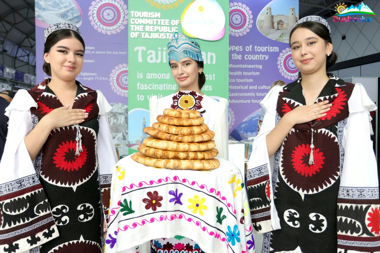 Three women in embroidered Tajik dress at the Tourism Committee’s stand, presenting a tower of traditional flatbread